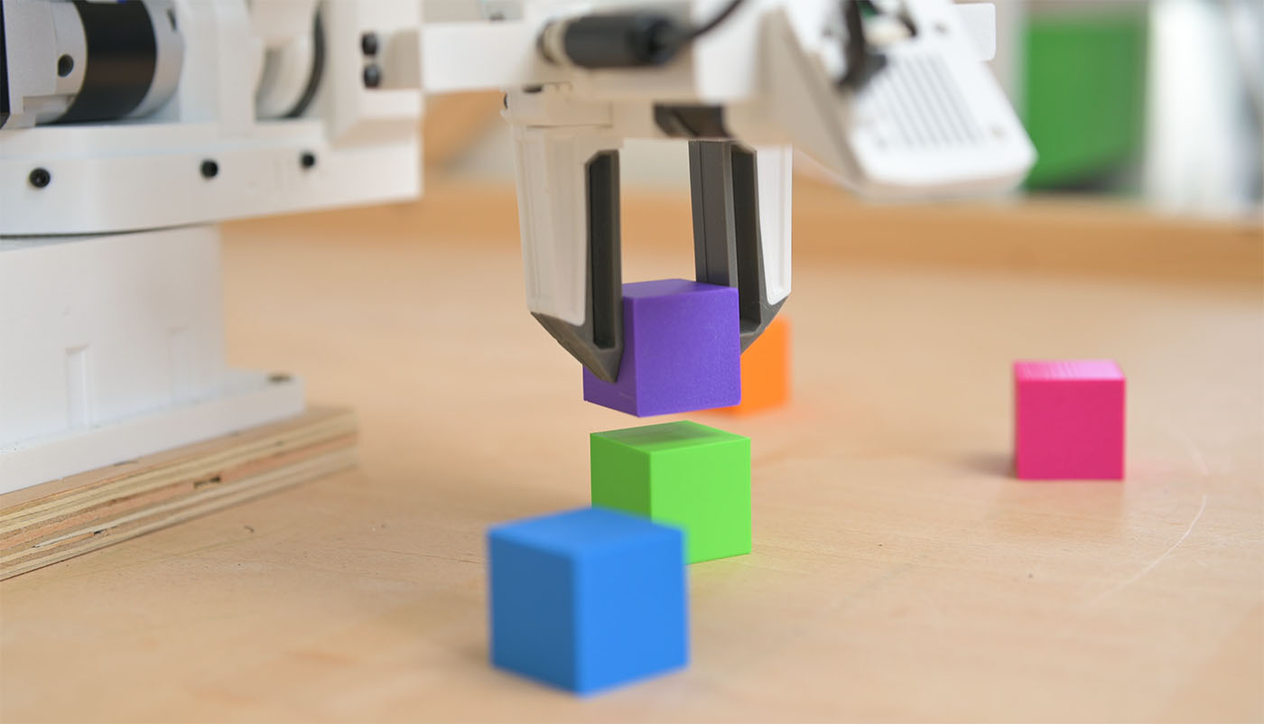 A robot gripper holding a purple block above a green one, with blue, orange and pink blocks on the bench